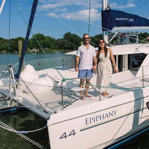 Couple smiling on a sailboat named Epiphany moored at a dock on a sunny day.