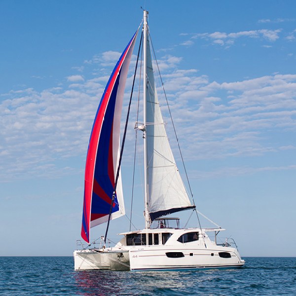 Catamaran sailing with red and blue spinnaker on a clear day at sea.