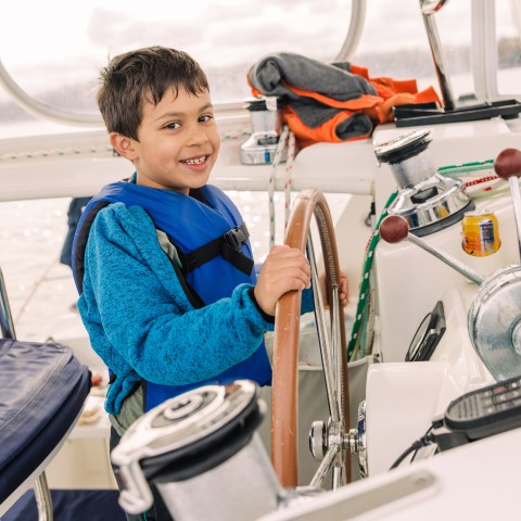 Smiling child wearing a life jacket steering a boat using a wooden wheel.