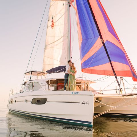 Couple standing on a sailing catamaran with colorful sails at sunset.